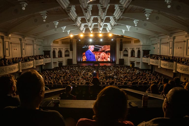 Packed theatre audience watches a performance on stage with large screen projection above the crowd.