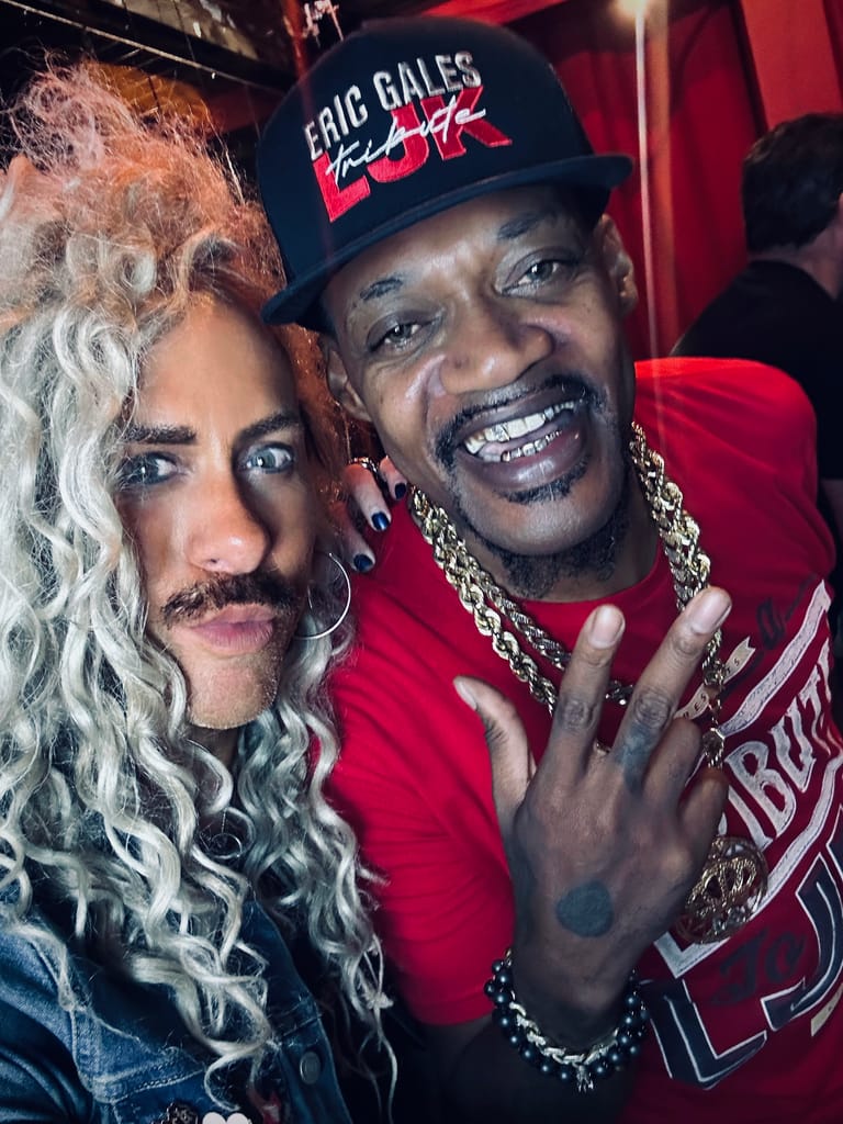 Two people pose together at a concert venue, one wearing an Eric Gales cap and red shirt with chain necklace, both smiling at the camera.