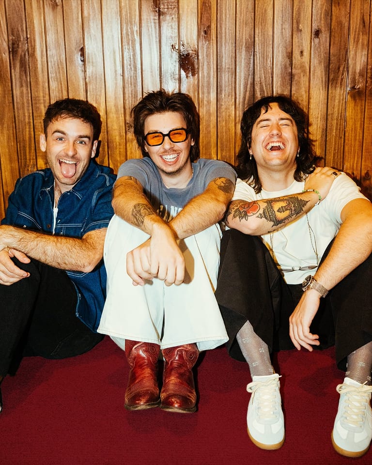 Three musicians with animated expressions pose together against a wooden backdrop, seated on a red surface.