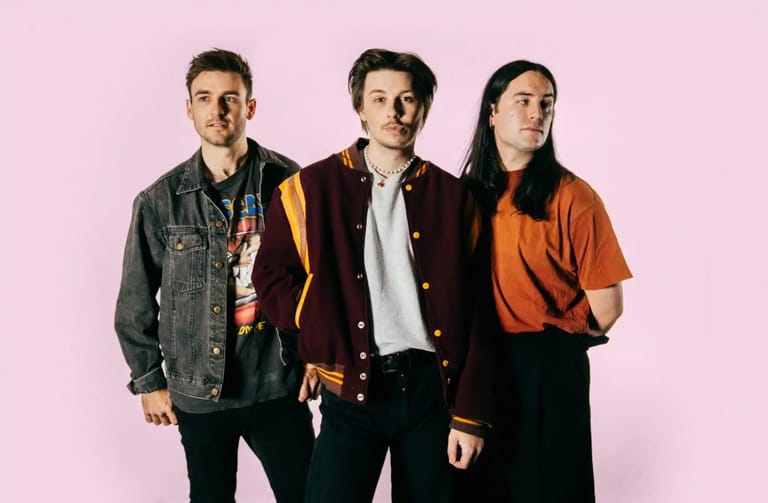 Three band members stand against a pink background wearing casual jackets and shirts.