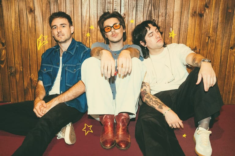 Three band members sit against wooden panelling, arms linked, wearing casual denim and vintage boots with star details.