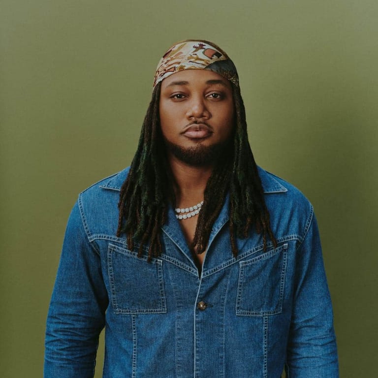 Person with long braids and patterned headwrap wearing denim jacket against green background.