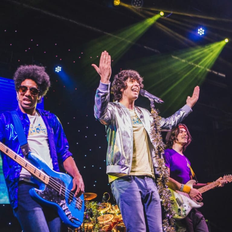 Three musicians perform on stage under yellow and blue spotlights, with the centre figure singing with arms raised.