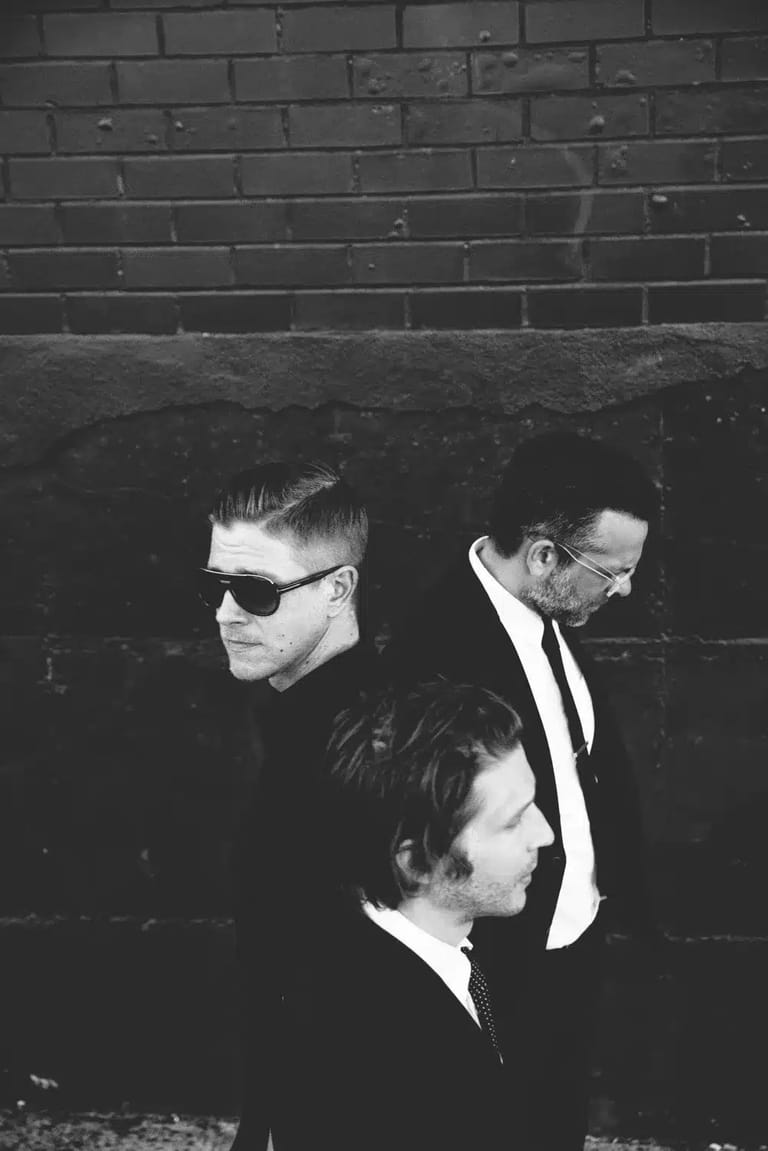Three men in dark suits and sunglasses stand against a brick wall in black and white.