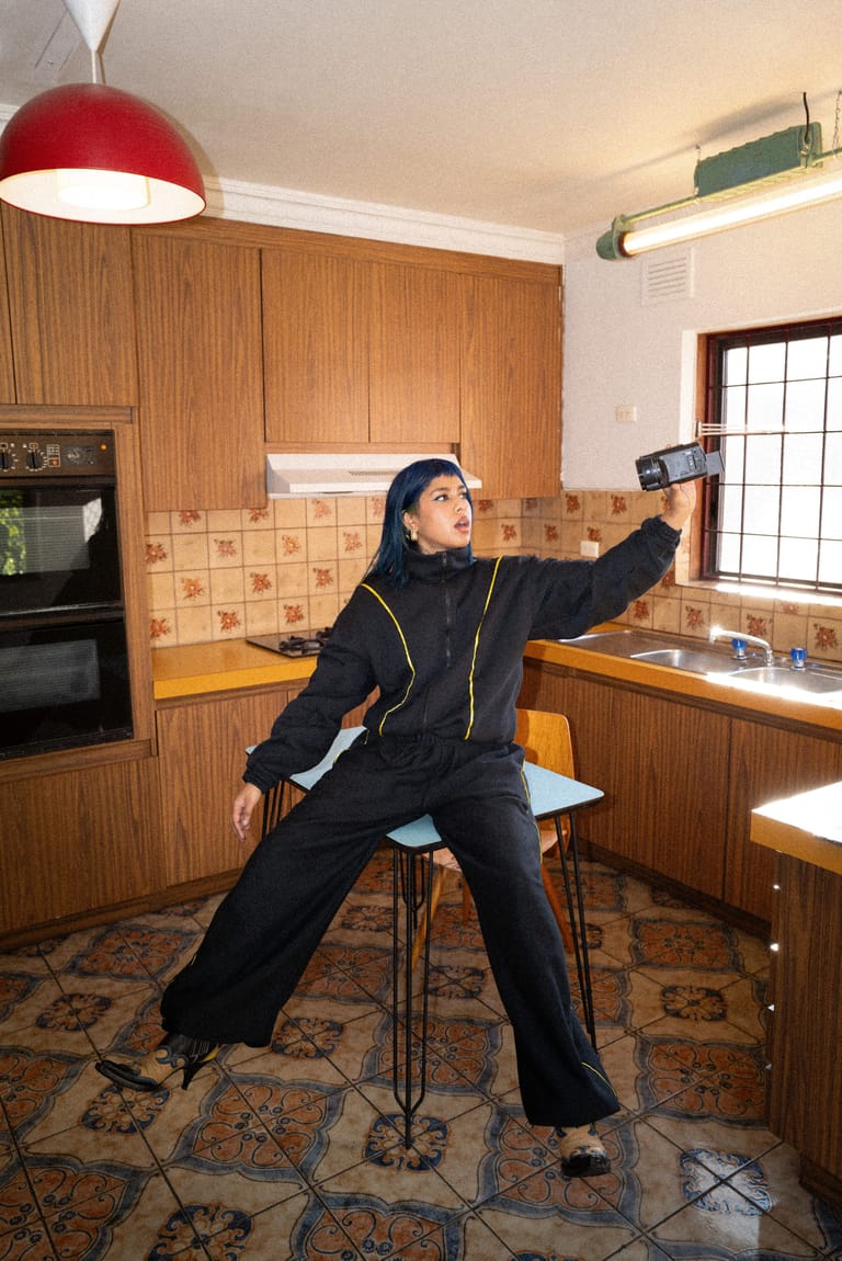 Person in black tracksuit striking a dynamic pose on a stool in a retro kitchen set, holding up a mobile device.