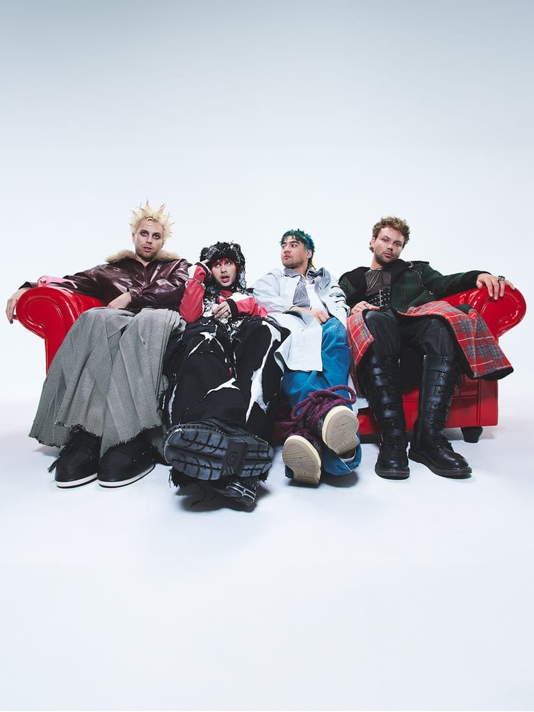 Four performers in bold, colourful styling sit together against a white background holding red fabric.