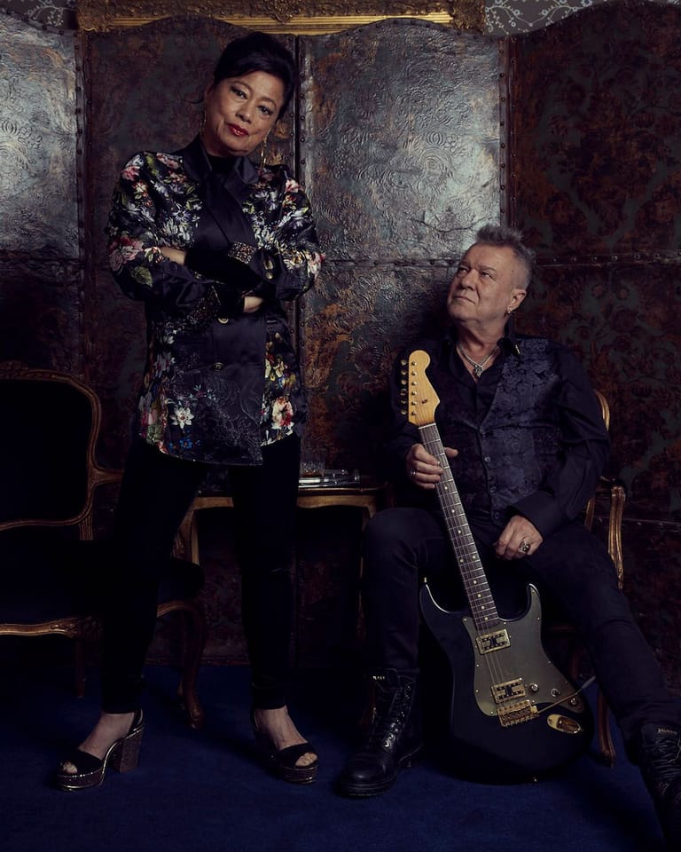 Two musicians pose against a weathered metal backdrop, one standing in a patterned jacket and one seated holding a bass guitar.