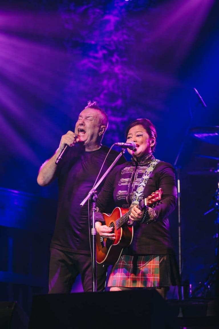 Two performers sing together on stage under purple and blue stage lighting, one holding a microphone and the other playing an acoustic guitar.