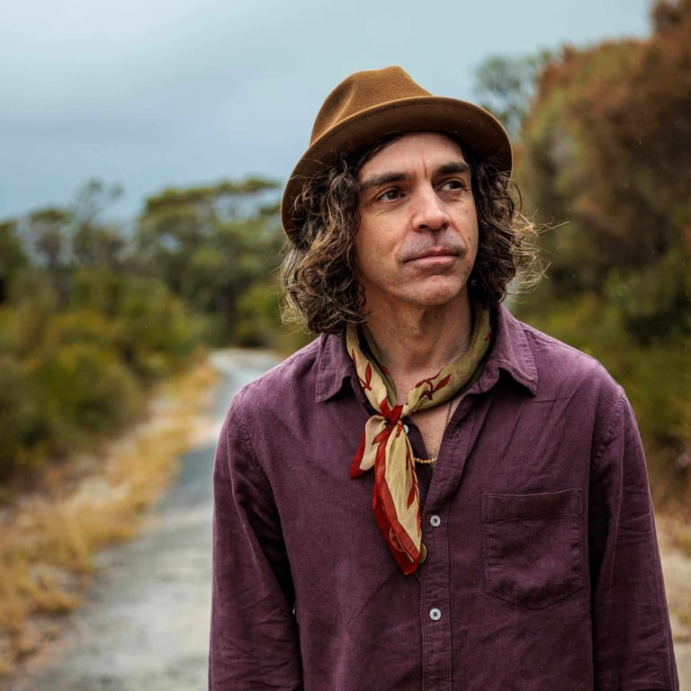 Man with long curly hair and brown hat stands on a rural road wearing a purple shirt and bandana.