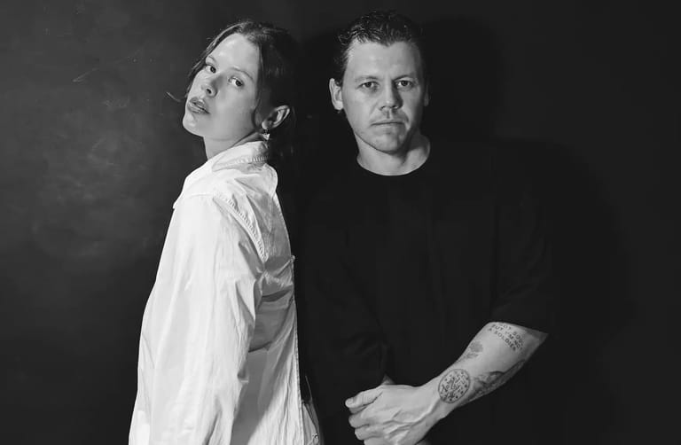 Two men pose back-to-back against a dark background in a black and white portrait.
