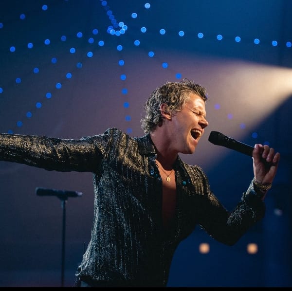 Singer in sparkly jacket performing at microphone against blue-lit stage backdrop with bokeh lights.