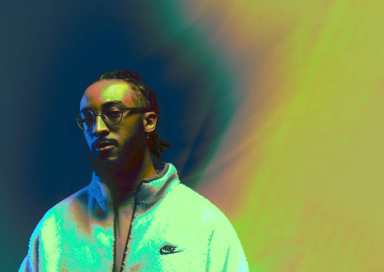 Person wearing sunglasses and a light jacket against a vibrant blue and yellow gradient background.
