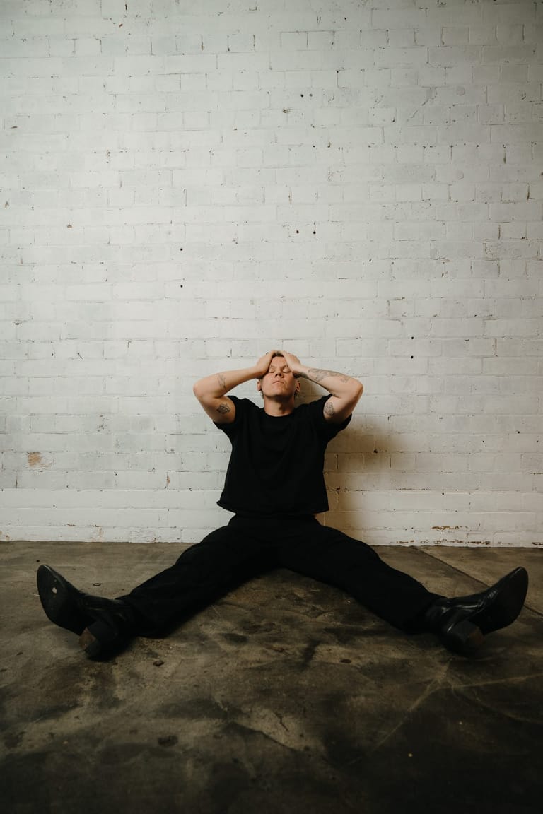 Person in black clothing sits with legs spread wide against a white brick wall, hands behind head in a relaxed pose.