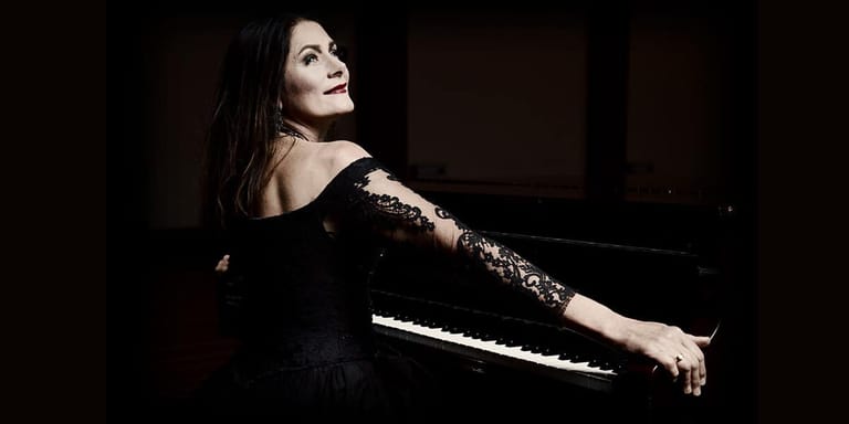 Woman in black off-shoulder dress with lace sleeves reclining against a piano in dramatic spotlight.