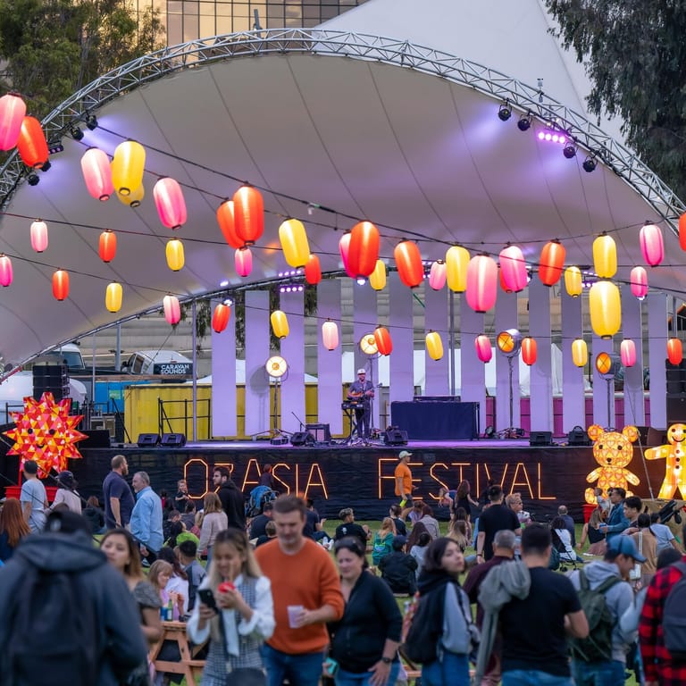 Colourful paper lanterns hang above an outdoor stage at a festival celebration with crowds gathered below.