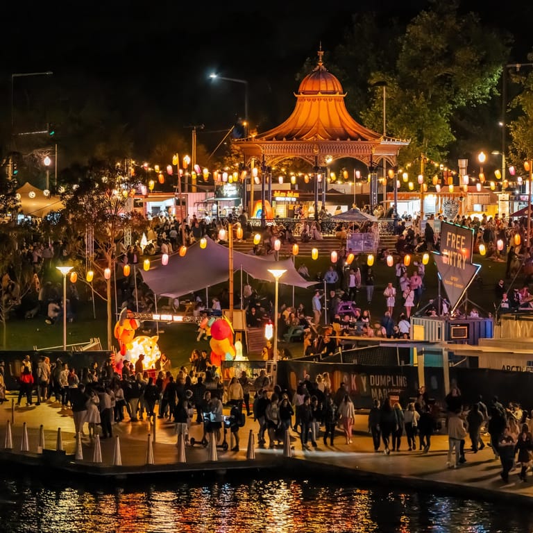 Crowds gather around an illuminated carousel and fairground rides at night beside a waterway.