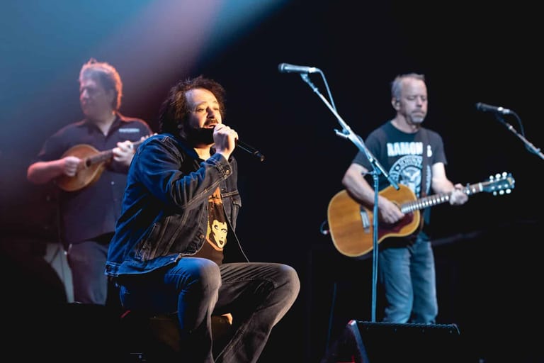 Three musicians perform on stage under coloured spotlights, with a vocalist centre stage and guitarists flanking either side.