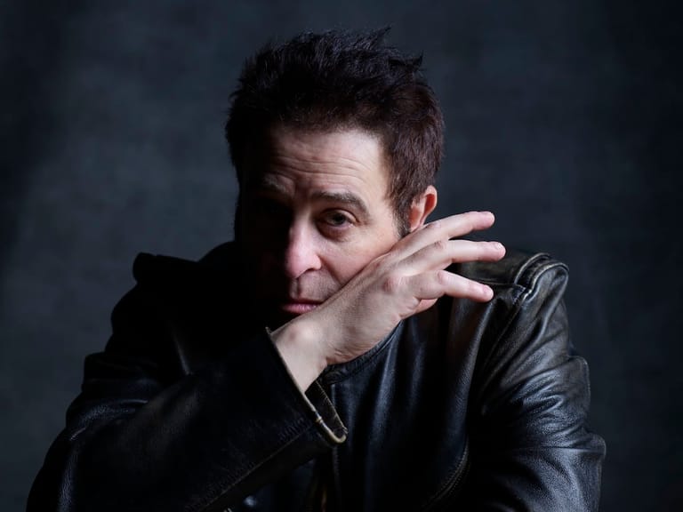 Man in black leather jacket gestures expressively against a dark background.