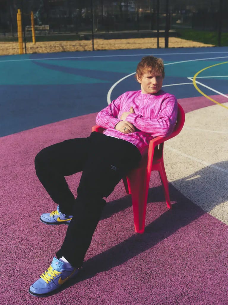 Person in pink jacket seated on red chair on colourful outdoor sports court in bright sunlight.