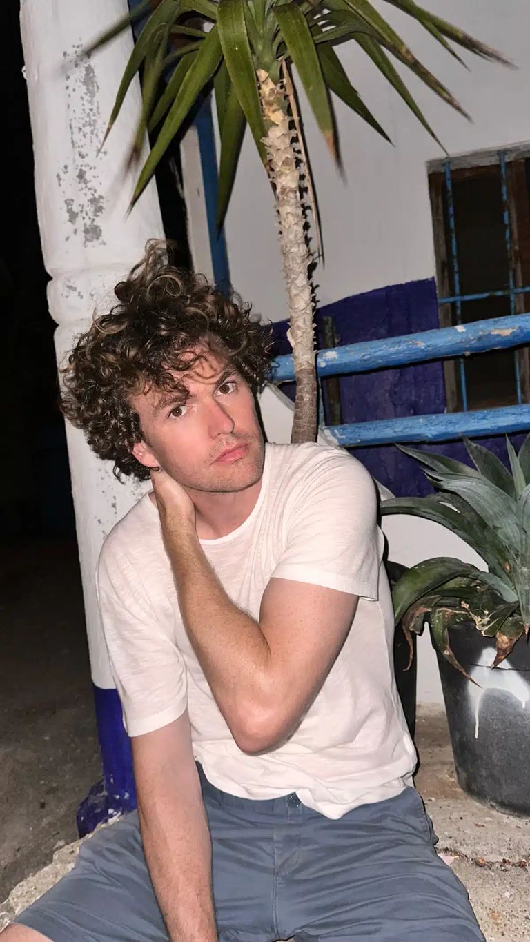 Person with curly dark hair in white t-shirt posing thoughtfully beside potted plants and blue shelving.