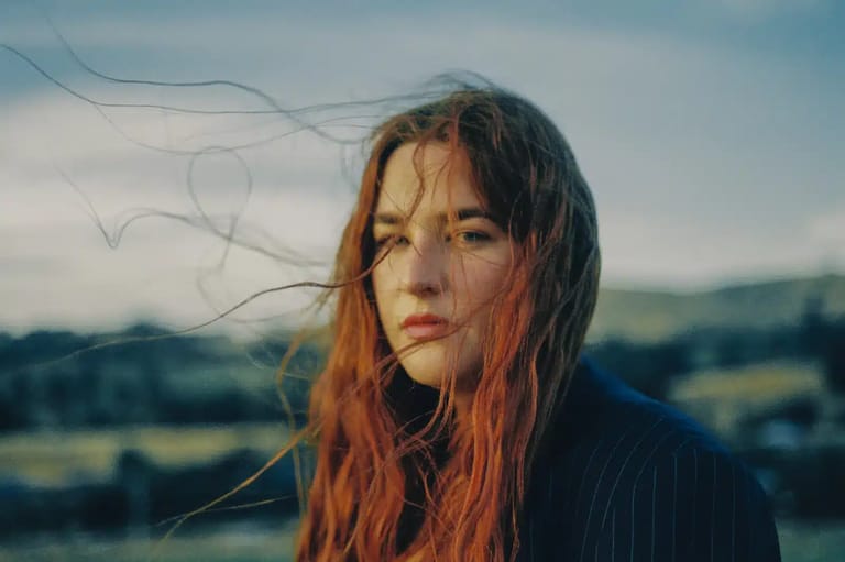 Person with long reddish-orange hair gazes toward camera against a hazy landscape backdrop.