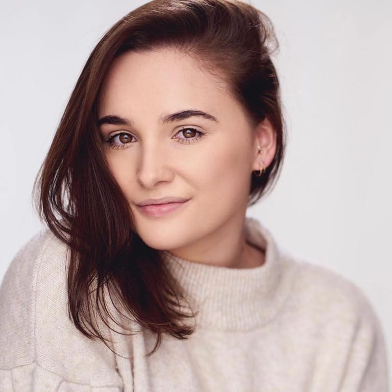 Woman with long reddish-brown hair wearing a cream knit sweater against a neutral background.
