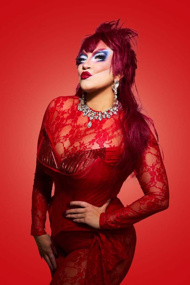 Performer in red lace dress and jewellery with bold makeup against bright red background.