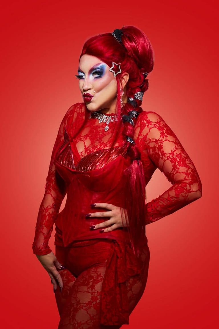Performer in striking red lace ensemble with bold makeup and styled hair against a vibrant red background.