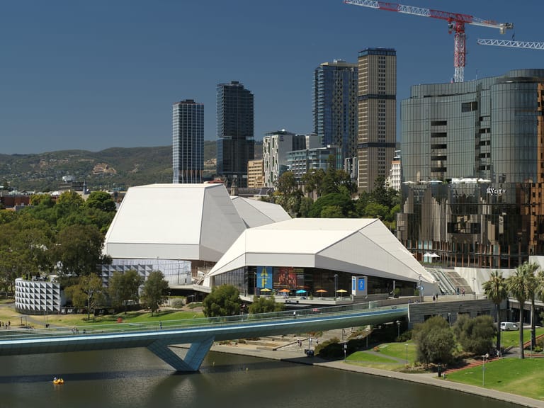 White angular contemporary building with blue bridge spanning river, Adelaide city skyline and hills beyond.