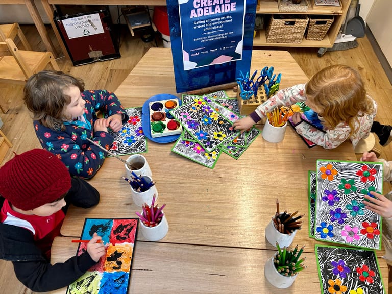 Two young children seated at a wooden table working on colourful art projects surrounded by paint, markers and craft supplies.