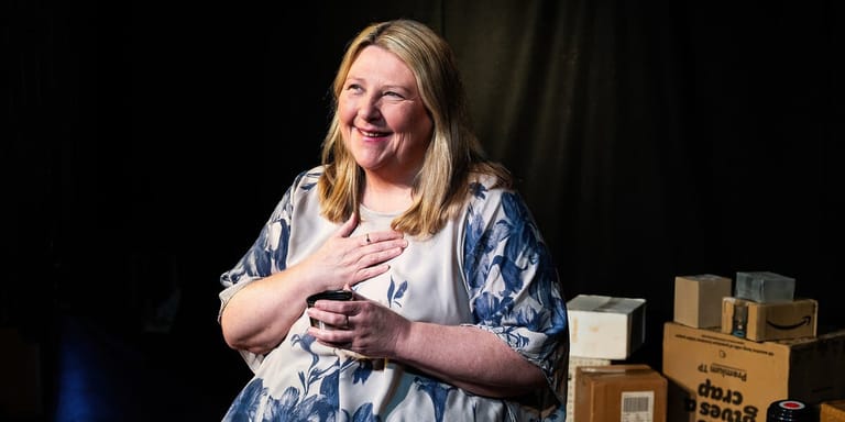 Woman with blonde hair smiling whilst speaking, hand on chest, against dark backdrop with cardboard boxes.