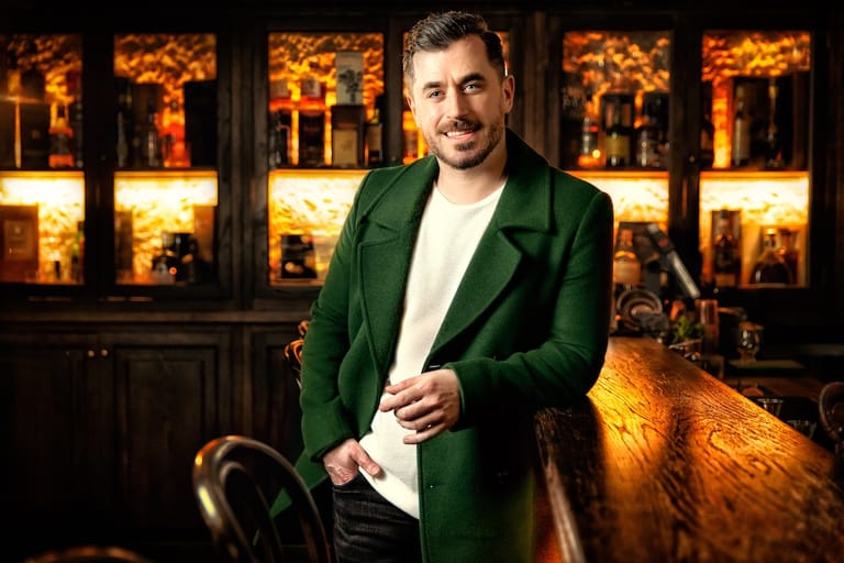 Man in green blazer and white shirt smiling at camera against warmly lit building facade.