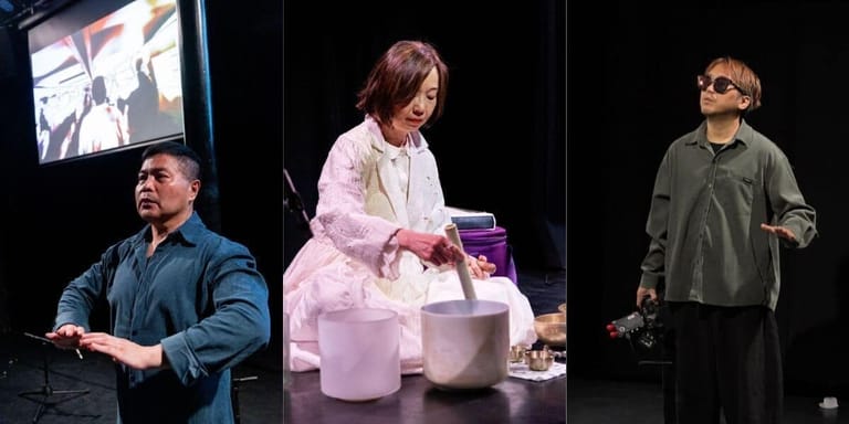 Three performers on stage during a theatrical production, with video screen and props visible in the background.