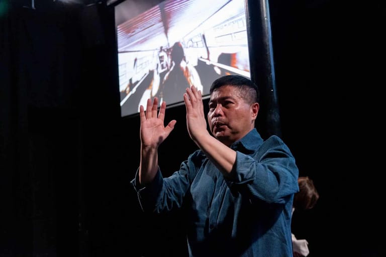 Man in denim shirt gestures expressively toward a large projected screen in a darkened performance space.