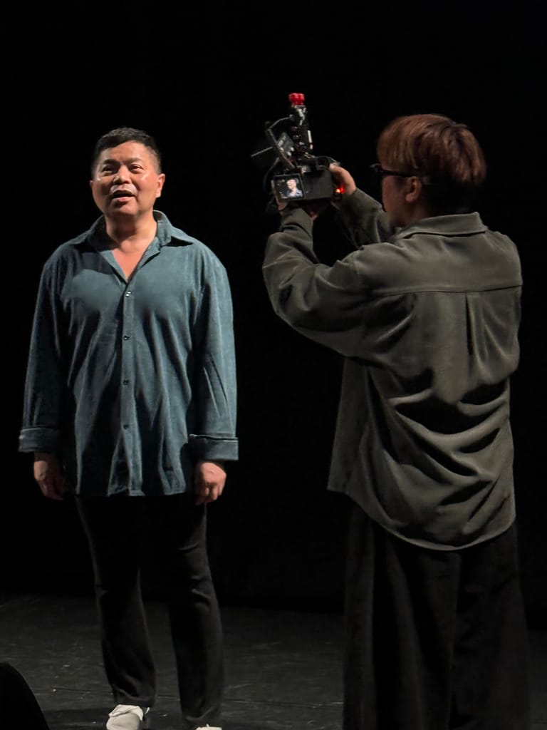 Filmmaker operating a camera interviews a man in a blue shirt on a darkened stage.