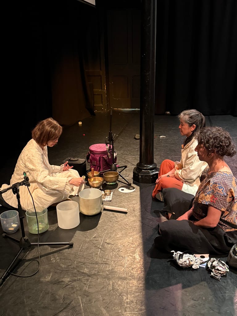 Three people seated on a theatre stage floor in conversation, with audio equipment and microphones visible around them.