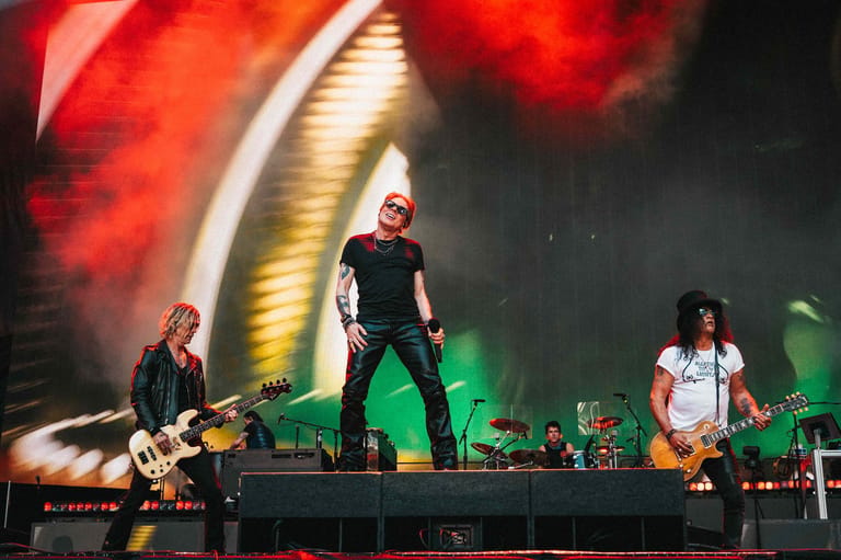 Band performing on stage with red and gold lighting, vocalist centre stage between two guitarists.