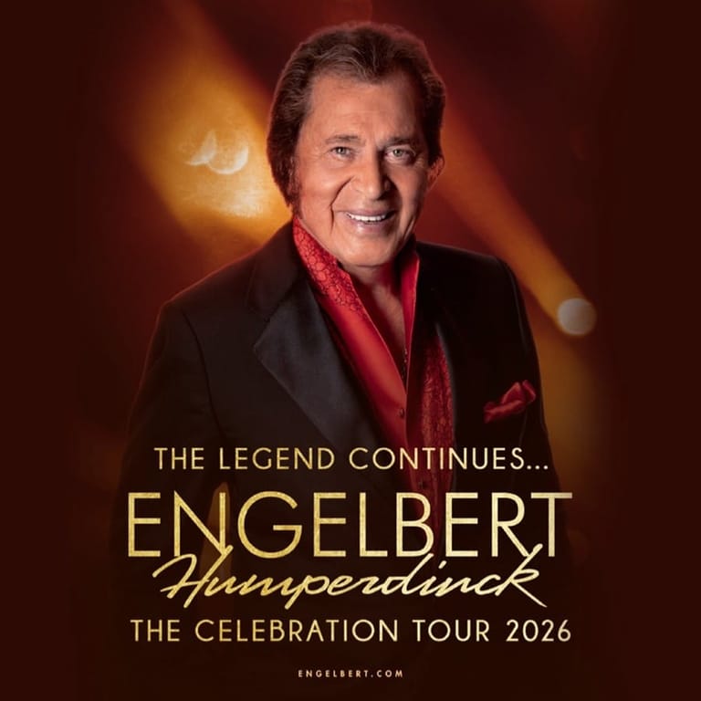 Man in black jacket and red shirt smiling against warm golden light with "The Legend Continues" tour text overlay.