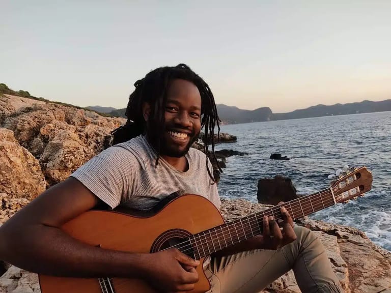 Musician holding an acoustic guitar on a rocky beach at sunset, water and distant hills visible behind.