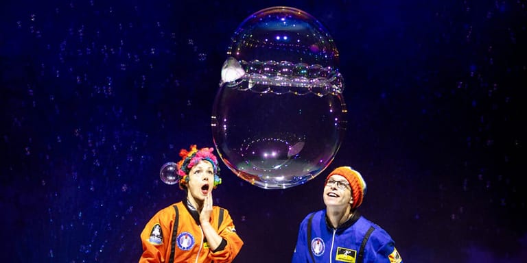 Two performers in colourful suits stand beneath a large illuminated bubble against a dark starry background.