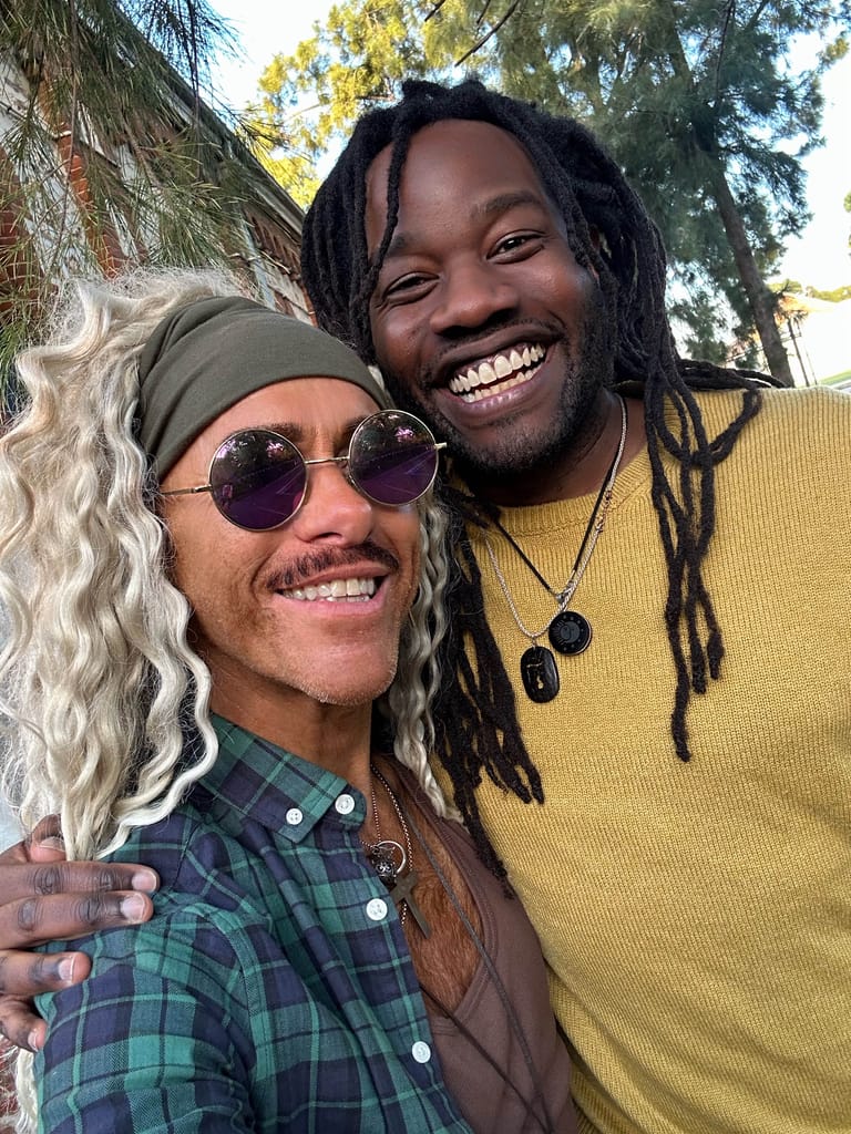 Two performers smile at the camera outdoors, one wearing a grey wig and purple sunglasses, the other in a yellow shirt with dreadlocks.