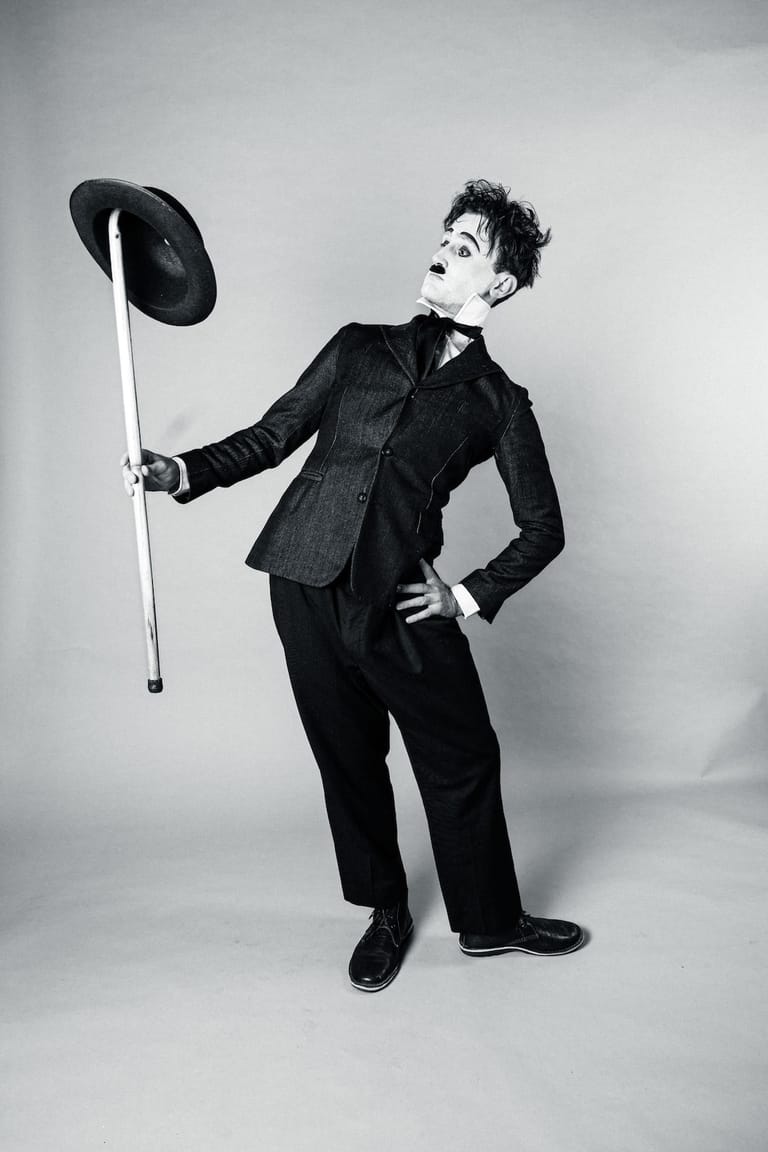Person in dark suit holding a cane with a hat, posed against neutral background in black and white studio portrait.