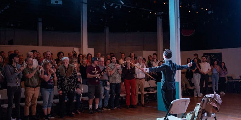 A speaker in dark clothing addresses a large gathered audience in an indoor venue at night.