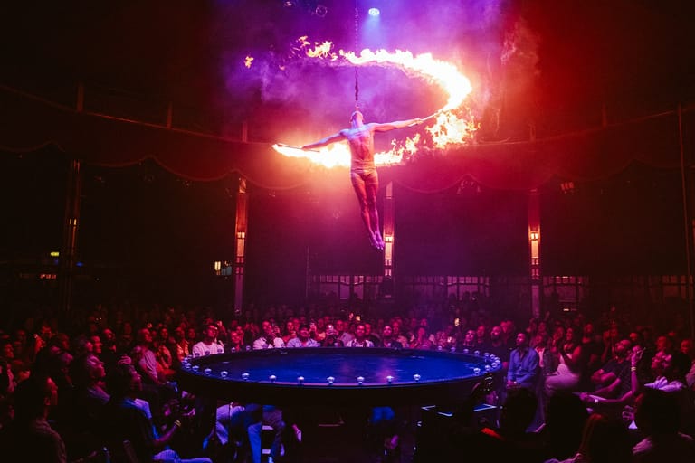 Fire performer holds flaming props above their head on a circular stage surrounded by audience members in purple and red lighting.