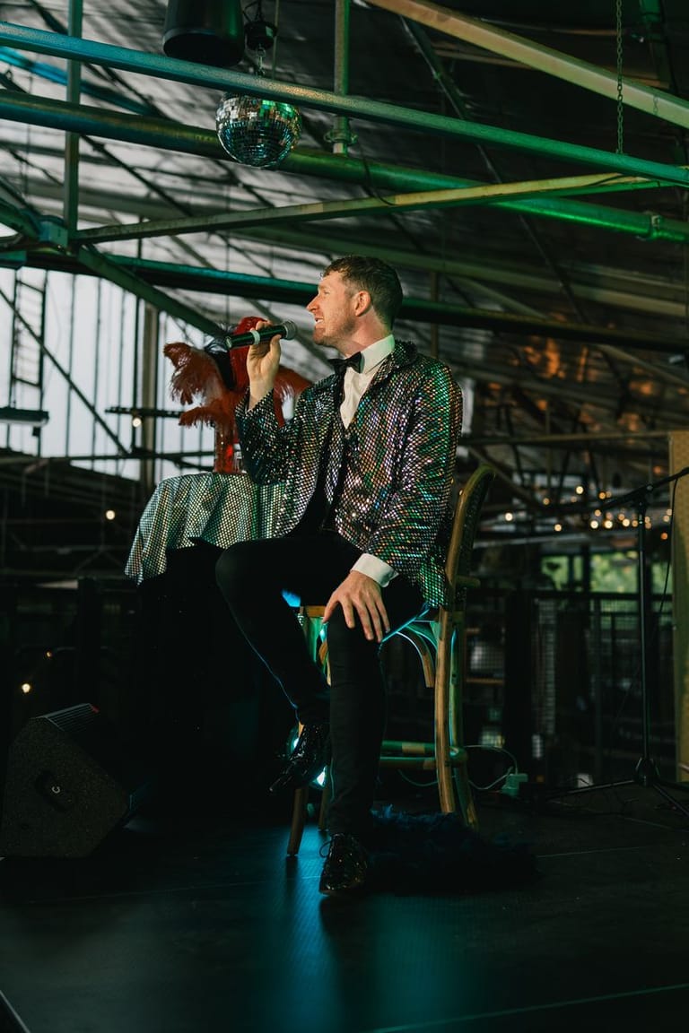 Man in a patterned green jacket sits holding a vintage puppet in an industrial warehouse setting.