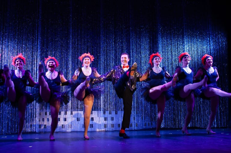 Seven performers in dark costumes dance synchronised on stage beneath blue theatrical lighting and a textured backdrop.