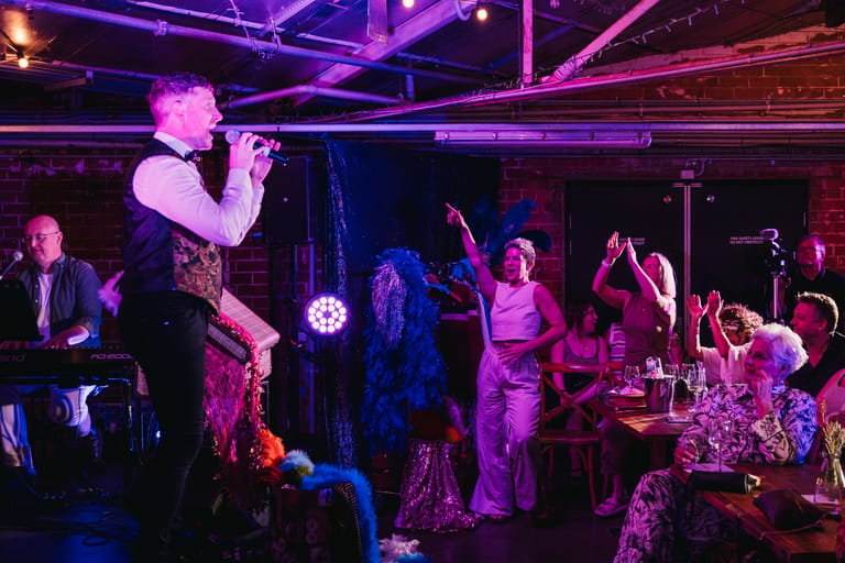 Performer singing into microphone centre stage with band and audience members under purple and red stage lighting.