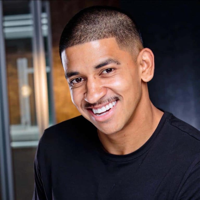 Smiling person in a black t-shirt against a dark interior backdrop with warm lighting.