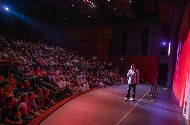 A performer stands alone on an illuminated stage before a large seated audience in a theatre lit by red and purple stage lights.