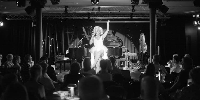 Performer in white clothing centre stage with arms raised, surrounded by audience members in a dimly lit venue.
